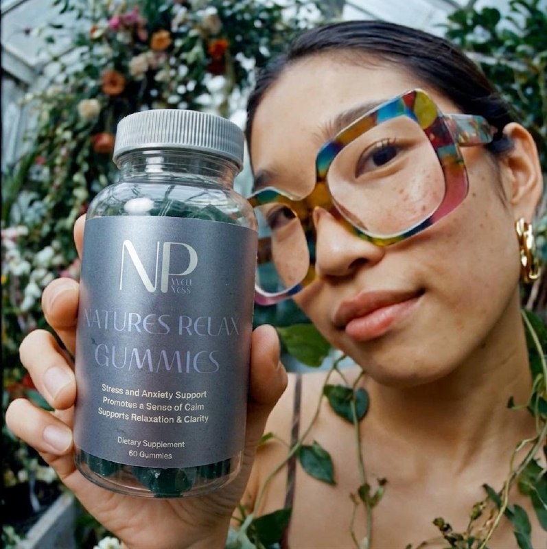 A woman in colourful glasses holding a bottle of Natures Relax Gummies in a greenhouse