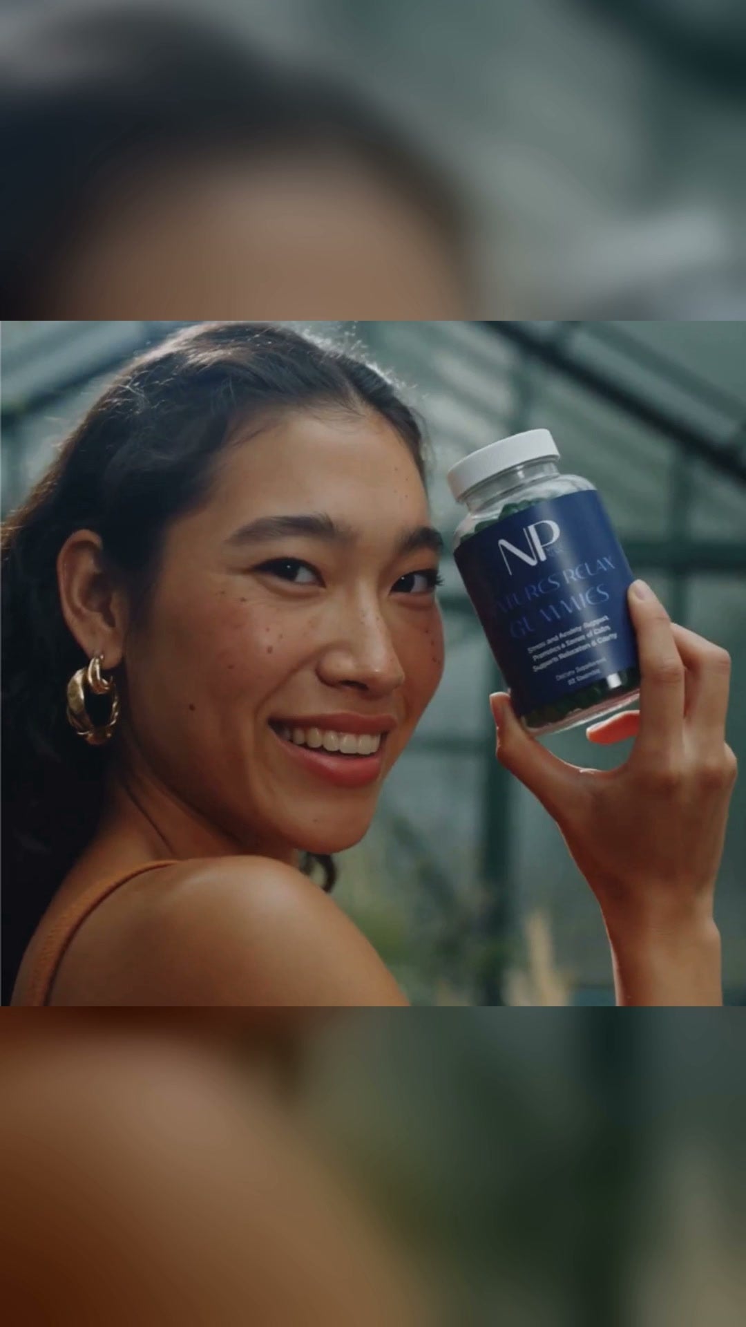 A woman crouched among wildflowers in a greenhouse, holding up a bottle of NP Nervous Relax Gummies