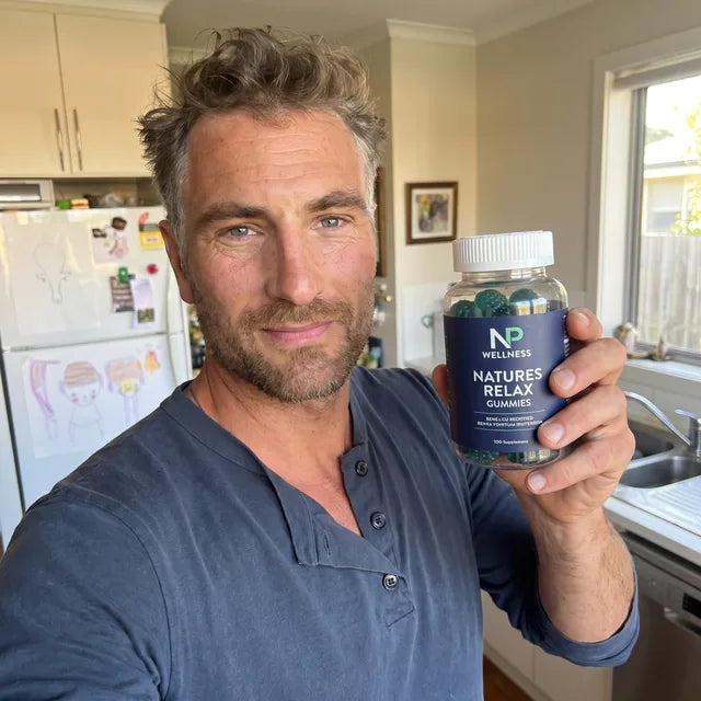 A dad in his kitchen holding up a bottle of Natures Relax Gummies while a toddler plays on the floor behind him